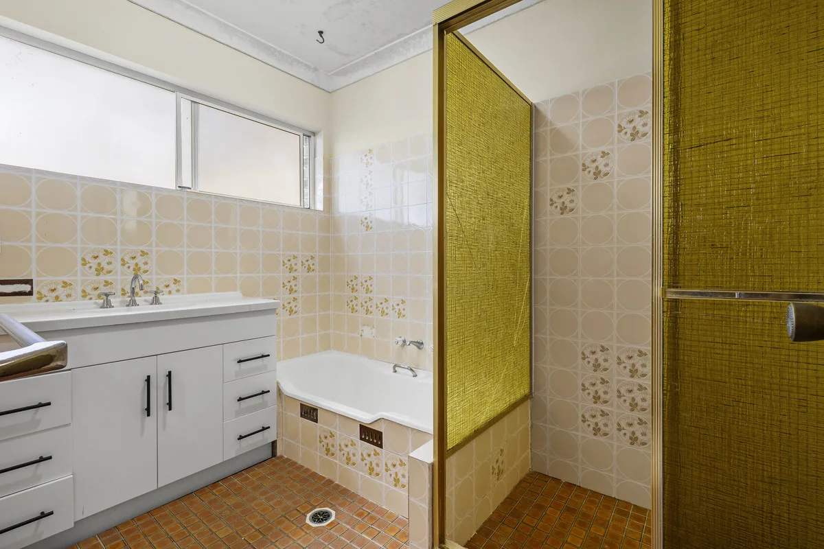 Dated bathroom with patterned wall tiles, a white vanity and a corner bath, before