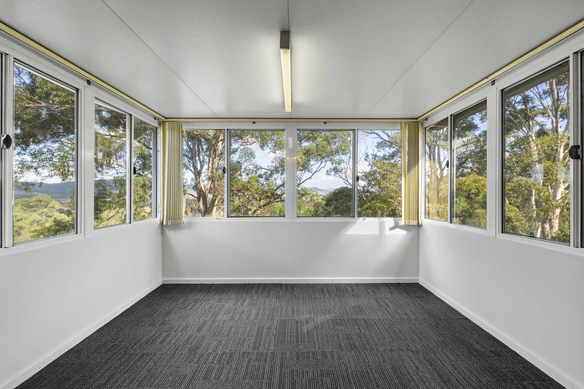 Empty enclosed sunroom with windows on three sides, grey carpet and treetop views, before